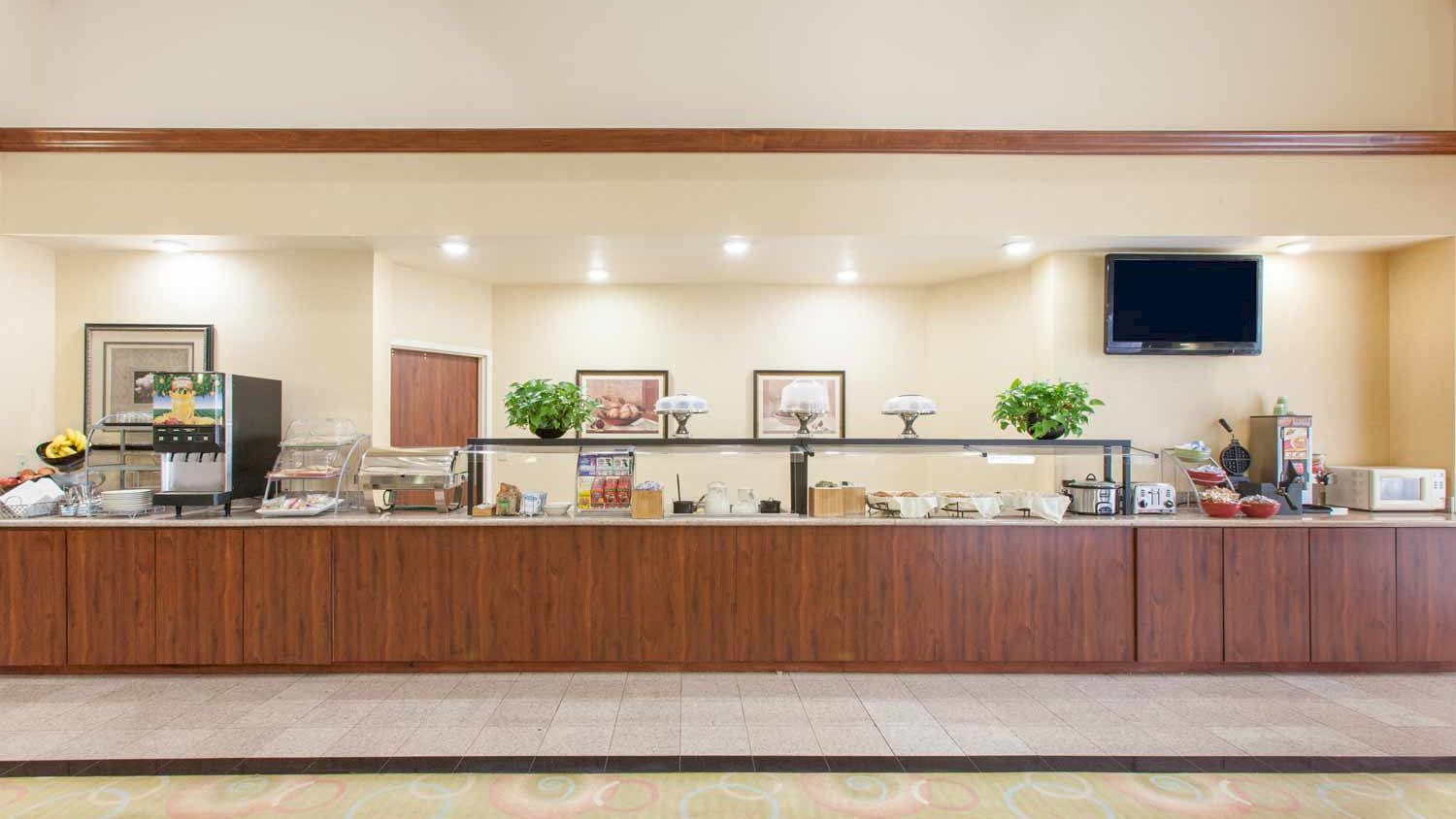 A self-service dining area with a food and drink counter, beverage dispensers, and plants on shelves in a clean, well-lit environment always ending the sentence.