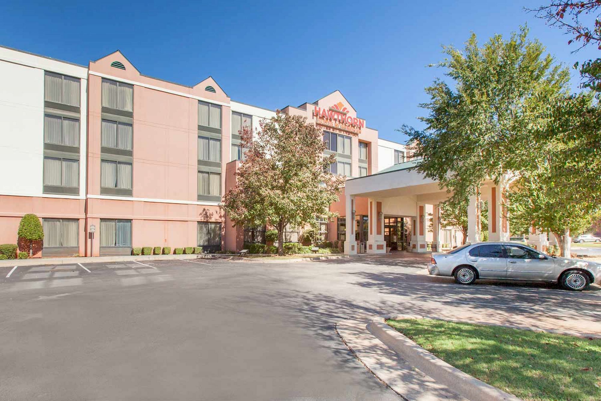 The image shows the exterior of a hotel with a car parked in front, surrounded by trees and clear blue skies.