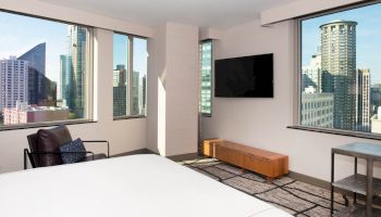 A modern hotel room with a large bed, a wall-mounted TV, a chair, and large windows offering a city skyline view ends the sentence.