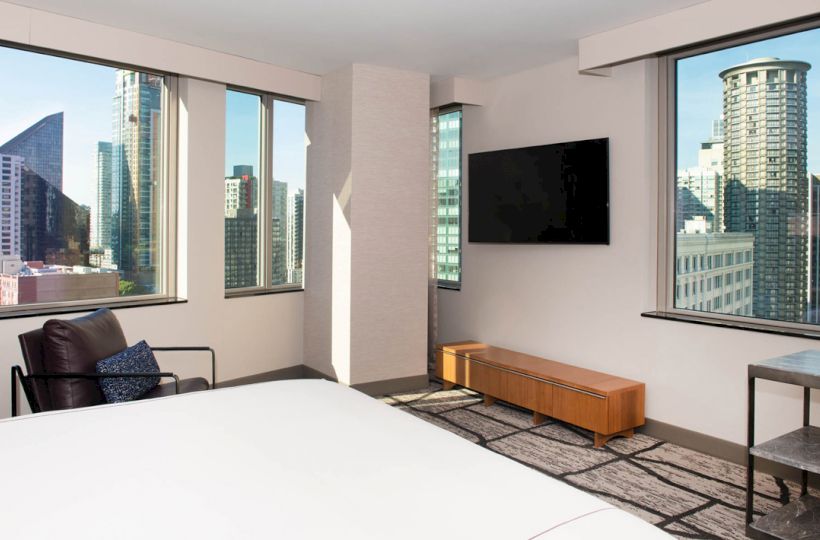 A modern hotel room with a large bed, a wall-mounted TV, a chair, and large windows offering a city skyline view ends the sentence.