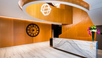 A modern lobby with a wooden spiral staircase, marble reception desk, black wall clock, and flower arrangement on the counter.