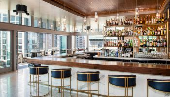 The image shows a modern bar with a sleek design, featuring a large bar counter, several bar stools, and a well-stocked liquor shelf.