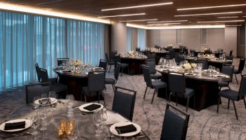 A banquet hall with round tables set with plates, glasses, and napkins, ready for an event; curtains and modern lighting complete the space.