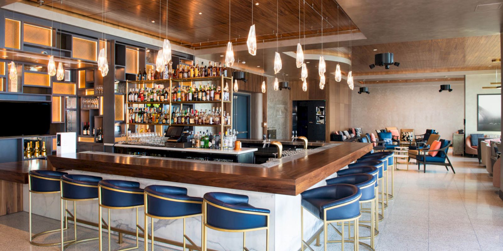 A modern bar with a wooden counter, blue barstools, and a display of various bottles. Pendant lights hang from the ceiling in this chic setting.