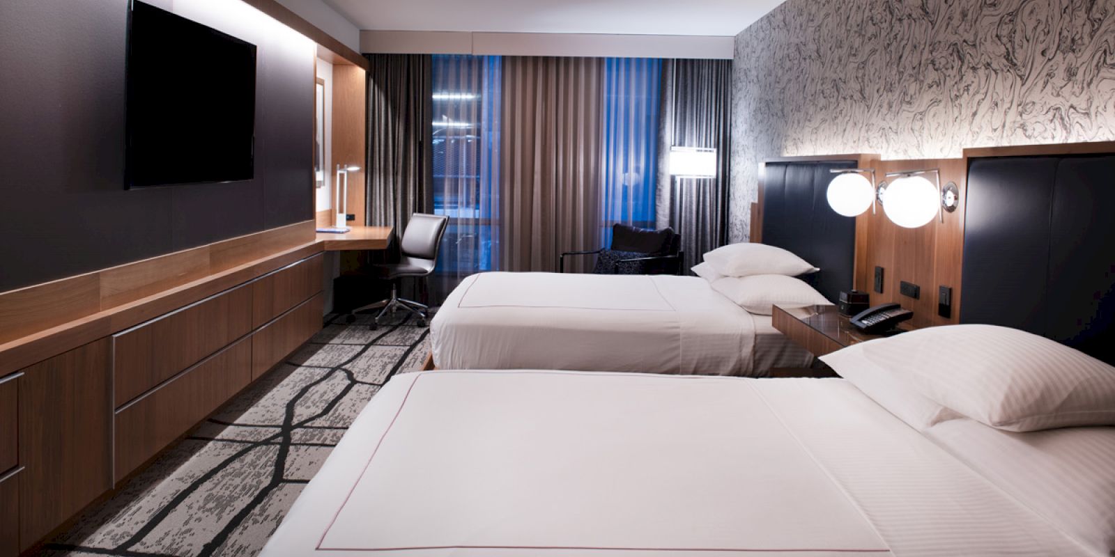 A modern hotel room features two double beds, a wall-mounted TV, a desk with a chair, and large windows with curtains, providing a welcoming ambiance.