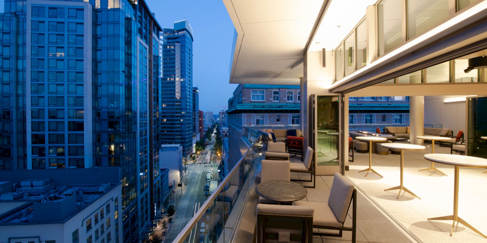 An urban balcony at twilight, with chairs and tables on one side, overlooking a street with tall buildings under a darkening blue sky.