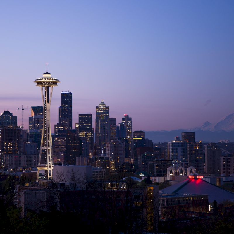 The image shows a nighttime cityscape of Seattle, featuring the Space Needle, various high-rise buildings, and Mount Rainier in the background.