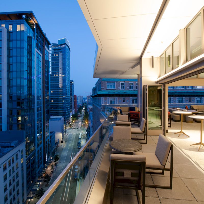 A modern balcony overlooking a cityscape with high-rise buildings. The area is furnished with tables and chairs, and the city lights are visible.