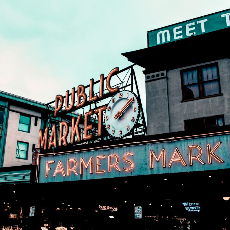 The image shows the entrance of a public market with a clock and signage reading "Farmers Market" in neon letters, surrounded by buildings.