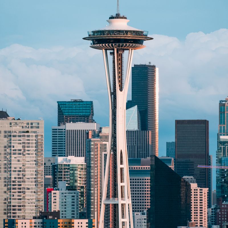 The image showcases the Space Needle tower with surrounding modern buildings in the city of Seattle, set against a backdrop of cloudy skies.