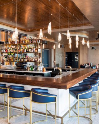 The image depicts a modern bar with blue bar stools, pendant lights, and a well-stocked counter in a spacious, elegantly designed setting.