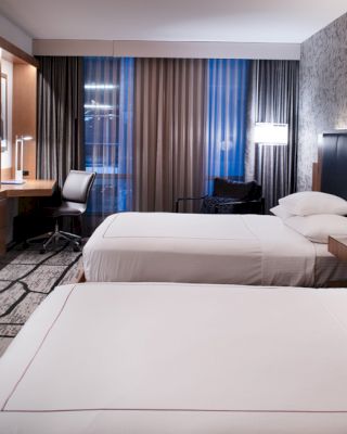 A modern hotel room with two double beds, a flat-screen TV, a desk, lamps, and large windows with curtains.