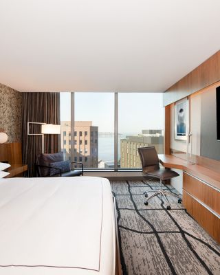 A modern hotel room with a king-sized bed, large flat-screen TV, desk with chair, a window with a city view, and contemporary decor on the walls.