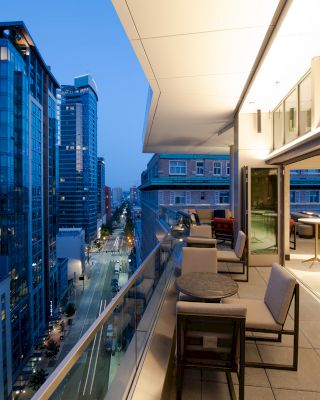 A modern balcony with seating offers a cityscape view of high-rise buildings and a lit street as dusk falls, creating a serene urban setting.