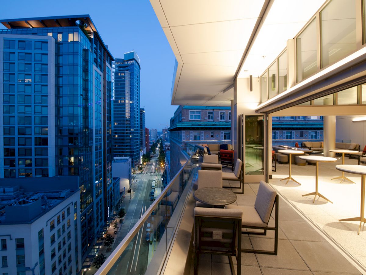 A modern cityscape view from a balcony with tables and chairs. The evening sky adds a serene contrast to the illuminated buildings.