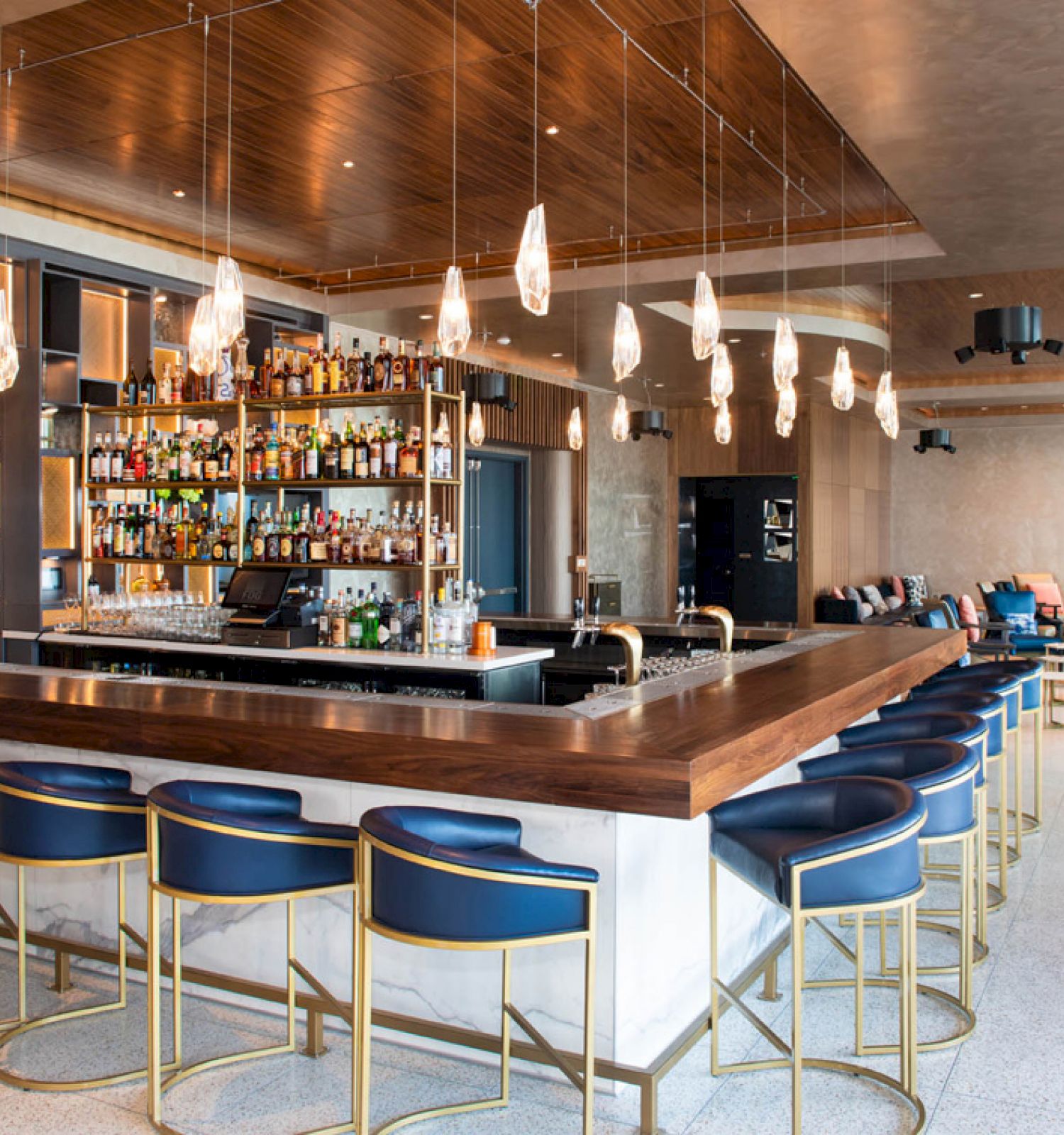 A modern bar with high stools around a wooden counter, illuminated by pendant lights, with shelves of bottles and a seating area in the background.