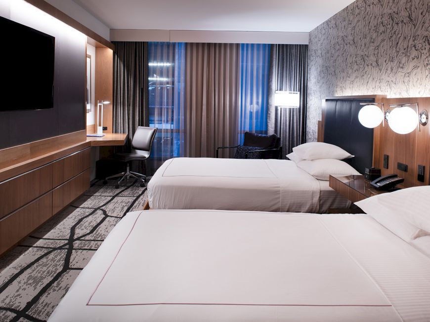 A modern hotel room with two neatly made beds, a wall-mounted TV, a work desk with a chair, and stylish decor with a patterned carpet.