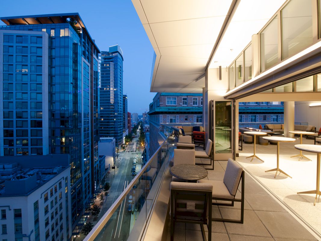 A modern cityscape view from a high-rise building's balcony with outdoor seating and tables, overlooking a street with more tall buildings.