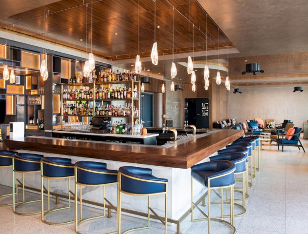 A modern bar with a large selection of alcohol, stylish blue bar stools, pendant lights, and cozy seating areas in a spacious, contemporary setting.