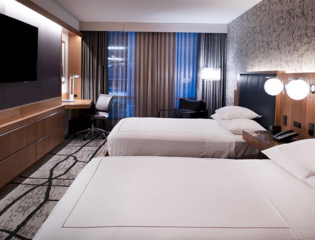 The image shows a modern hotel room with two double beds, a flat-screen TV, a desk with a chair, and stylish decor with a patterned wall.