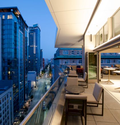 The image shows a cityscape with high-rise buildings at dusk, viewed from a modern balcony with seating and tables, adjacent to an indoor space.