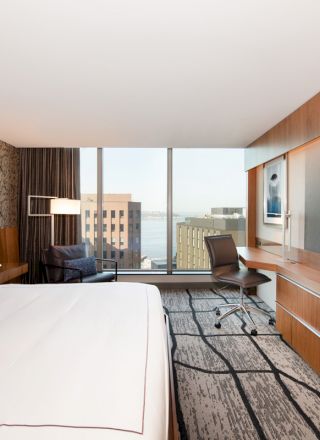 A modern hotel room with a large bed, desk, chair, TV, floor-to-ceiling window, and city view in the background.