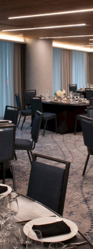  This image shows an elegantly set dining area with round tables, black chairs, and dinnerware, under modern lighting and near large curtained windows.