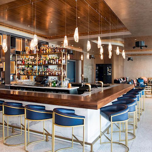 The image shows a modern bar with a lengthy counter, stylish blue and gold bar stools, a well-stocked liquor display, and ambient lighting.