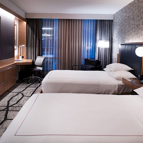 The image shows a modern hotel room with two neatly made beds, a flat-screen TV, a desk with a chair, and large windows with curtains.