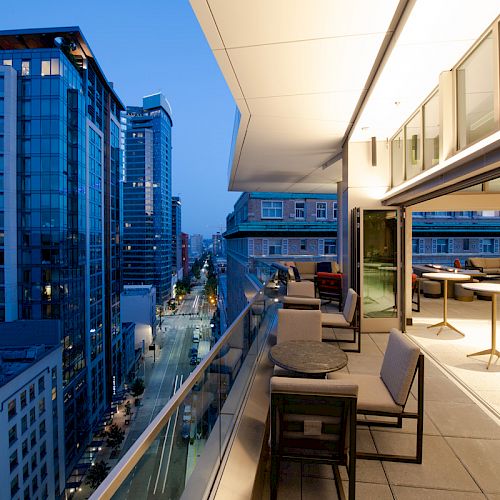 A modern urban balcony with seating overlooks a city street and adjacent skyscrapers at dusk, with indoor and outdoor areas visible.