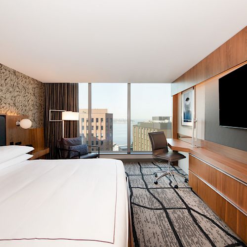A modern hotel room with a large bed, wall-mounted TV, desk, chair, and large window offering a view of buildings and water in the distance.