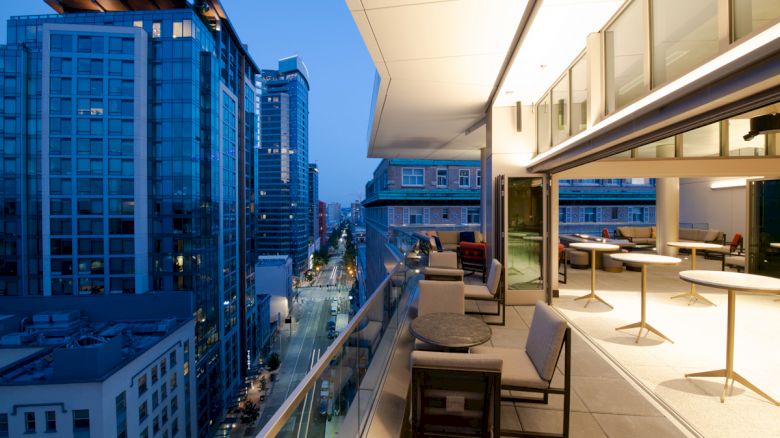 The image shows a modern balcony with seating and tables, overlooking a city street lined with tall buildings at dusk.