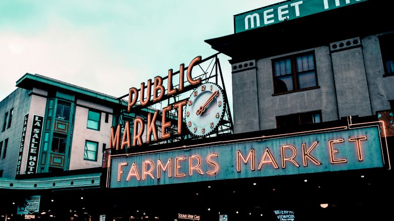 A vibrant public market with neon signs reading "Public Market" and "Farmers Market." The clock shows the time, and nearby buildings are visible.