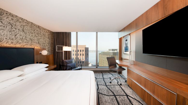 A modern hotel room with a large bed, a desk with a chair, a wall-mounted TV, and a window with a city view.