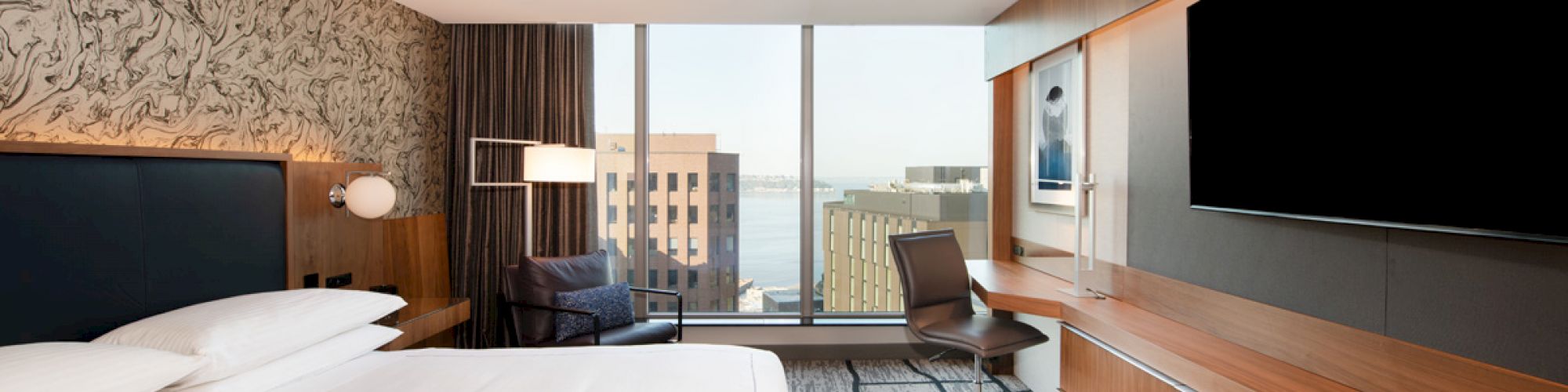 A modern hotel room with a large bed, desk, chair, flat-screen TV, and a window with a cityscape view.