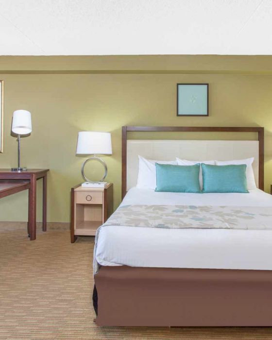 A neatly organized hotel room featuring a bed, two bedside tables with lamps, a desk with a chair, and framed artwork on the wall.