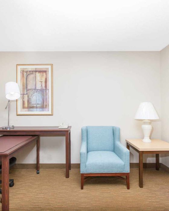 The image shows a small office with a desk, chair, lamp, artwork, an armchair, and a side table with a lamp. The decor is minimal and modern.